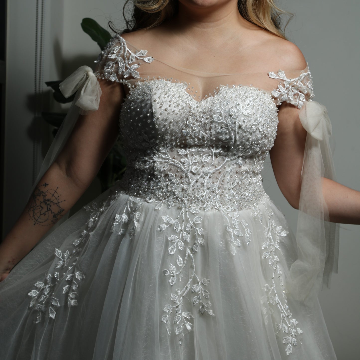 This is a light, ethereal wedding dress featuring delicate floral embroidery and beadwork. The gown has an off-the-shoulder design with sheer tulle straps tied into bows, adding a romantic touch. The bodice is adorned with intricate pearl and lace detailing, while the flowing tulle skirt is embellished with embroidered leaves, creating an elegant and whimsical look.