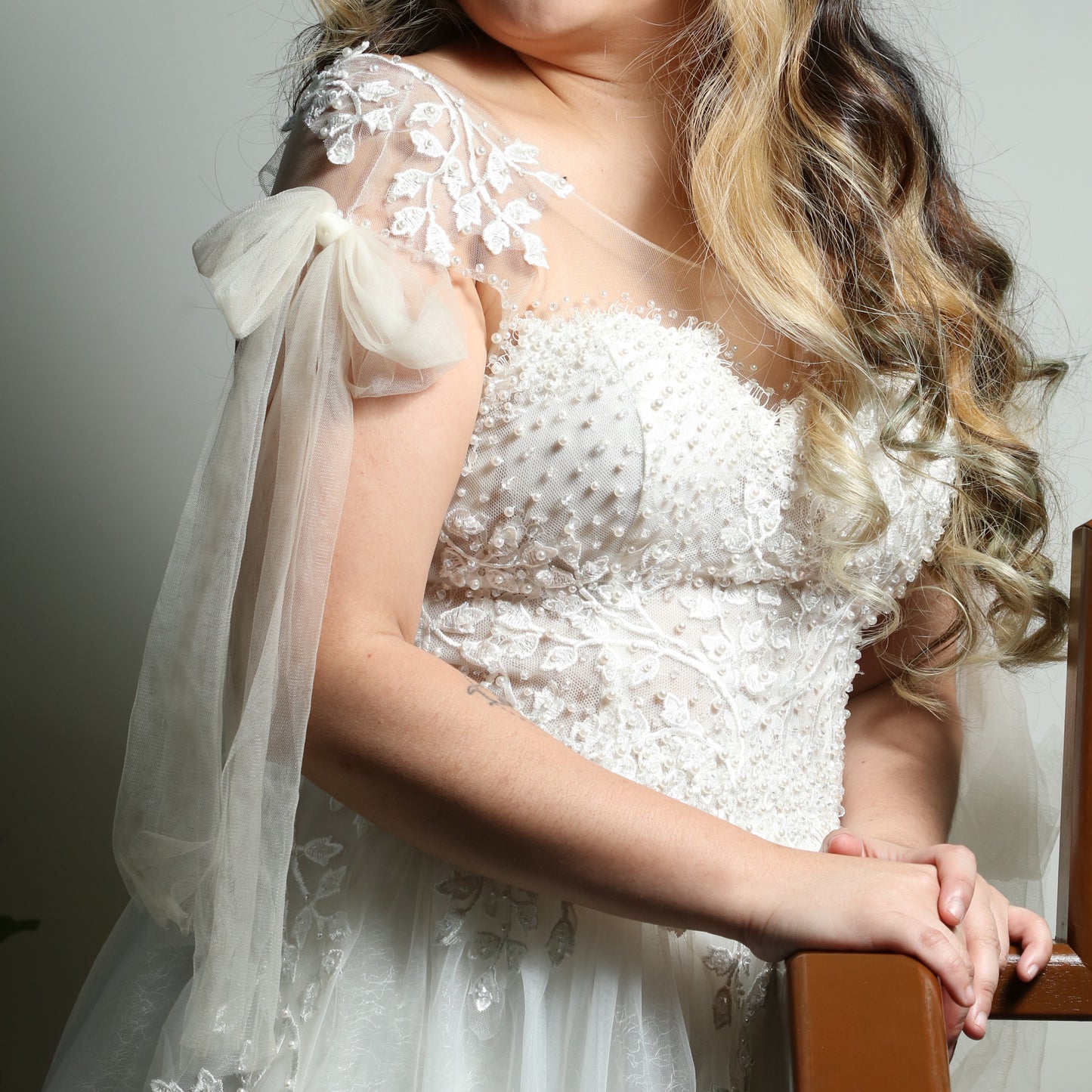 This is a light, ethereal wedding dress featuring delicate floral embroidery and beadwork. The gown has an off-the-shoulder design with sheer tulle straps tied into bows, adding a romantic touch. The bodice is adorned with intricate pearl and lace detailing, while the flowing tulle skirt is embellished with embroidered leaves, creating an elegant and whimsical look.