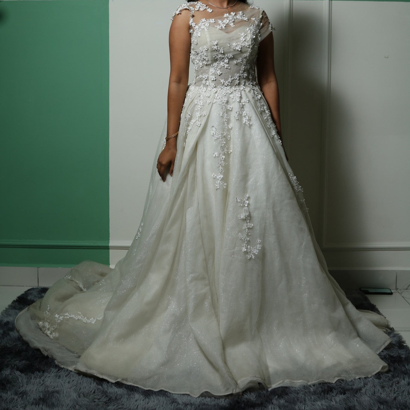 This gown beautifully merges modern elegance with delicate detailing. The base features a sleek, fitted tube-style inner dress that contours the body, creating a sophisticated silhouette. Over this, an ethereal layer of sheer fabric is adorned with intricate floral lace appliqués, adding a romantic and whimsical touch. The asymmetrical illusion neckline enhances the design, blending seamlessly with the floral embroidery for a soft yet striking effect.