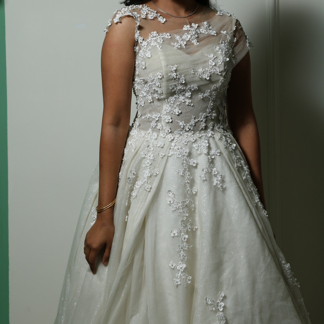 This gown beautifully merges modern elegance with delicate detailing. The base features a sleek, fitted tube-style inner dress that contours the body, creating a sophisticated silhouette. Over this, an ethereal layer of sheer fabric is adorned with intricate floral lace appliqués, adding a romantic and whimsical touch. The asymmetrical illusion neckline enhances the design, blending seamlessly with the floral embroidery for a soft yet striking effect.