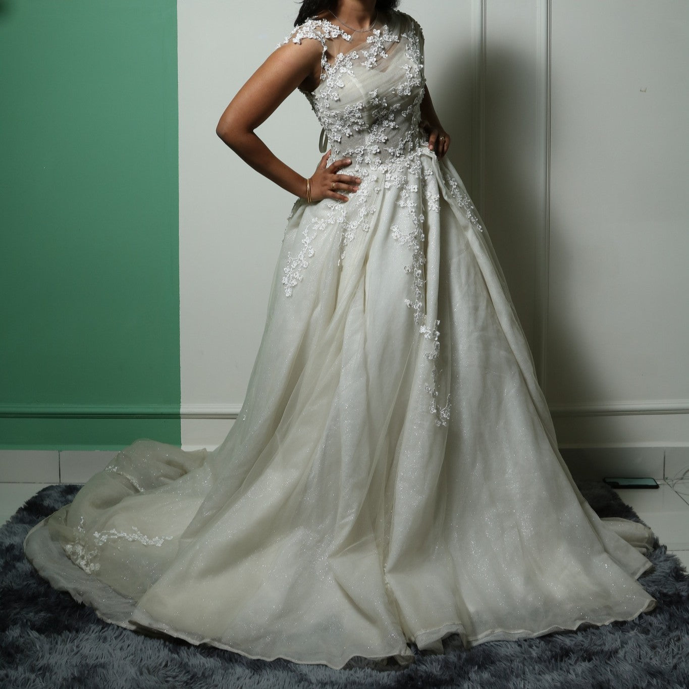 This gown beautifully merges modern elegance with delicate detailing. The base features a sleek, fitted tube-style inner dress that contours the body, creating a sophisticated silhouette. Over this, an ethereal layer of sheer fabric is adorned with intricate floral lace appliqués, adding a romantic and whimsical touch. The asymmetrical illusion neckline enhances the design, blending seamlessly with the floral embroidery for a soft yet striking effect.