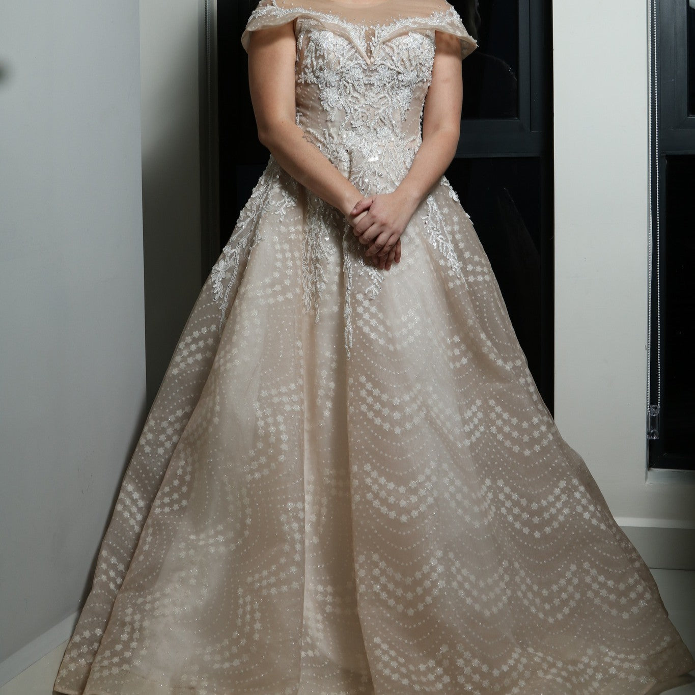 The dress in the image is an elegant, off-the-shoulder ball gown with intricate lace and bead detailing. The bodice features delicate embroidery that extends into the voluminous skirt, which is adorned with a subtle pattern of sparkling embellishments. The gown has a structured, layered design, making it visually striking but also quite heavy. The soft champagne or blush tone adds a romantic and timeless touch to the overall look.