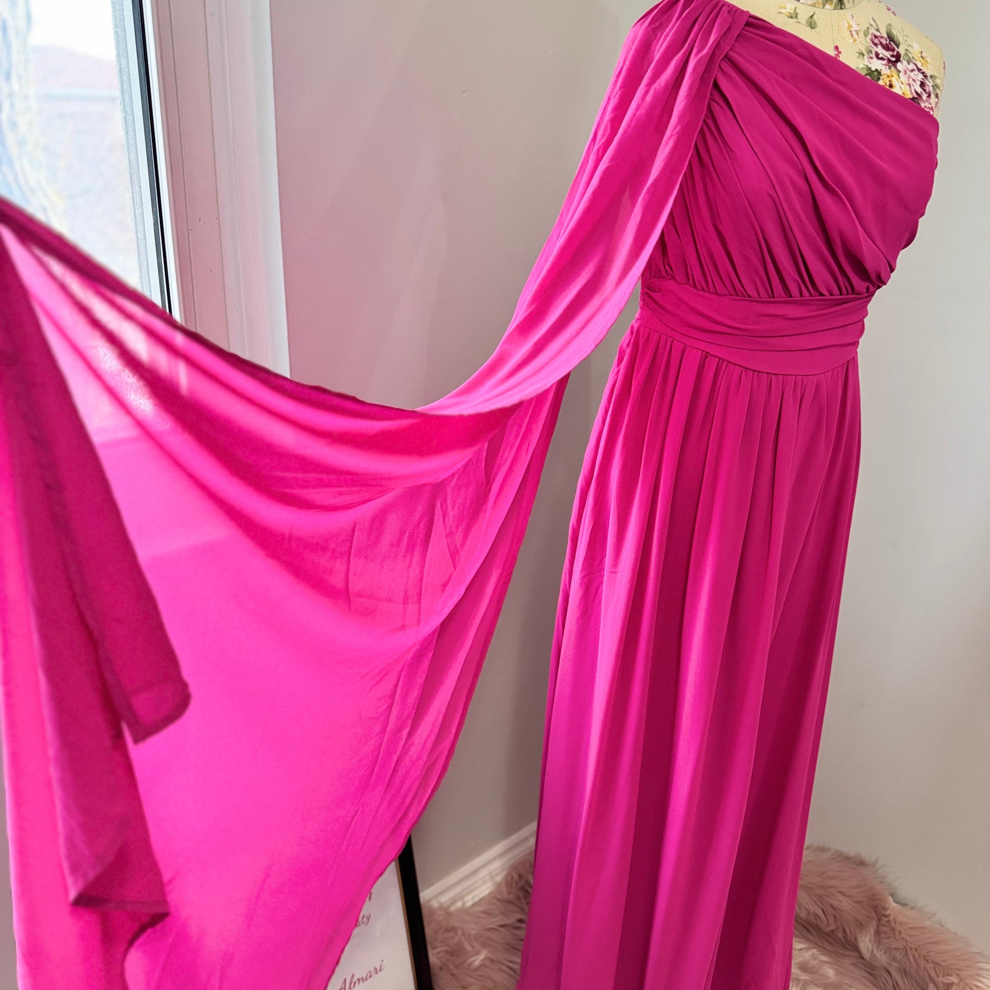 Step into effortless elegance with this breathtaking fuchsia one-shoulder gown, designed to make a statement at any event. Featuring a gracefully draped sleeve that cascades like a flowing waterfall, this dress exudes confidence, sophistication, and timeless charm. The cinched waist enhances your silhouette, while the soft, flowing fabric moves beautifully with every step.