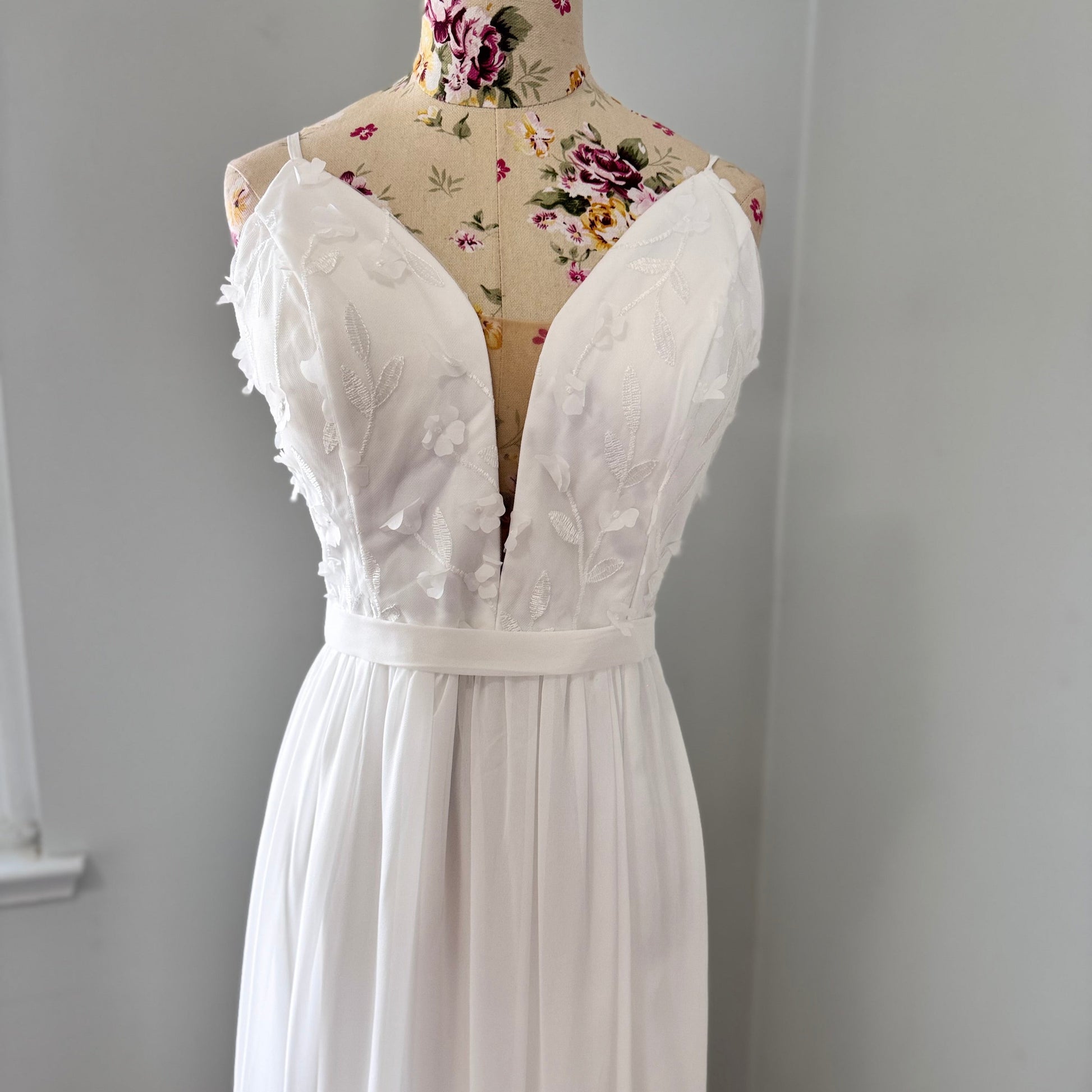 Elevate your style with this breathtaking white gown, designed to captivate with its effortless elegance and timeless charm. Featuring a flowy A-line silhouette, this dress showcases a delicate plunging V-neckline adorned with intricate 3D floral appliqués and embroidered leaf details, adding a soft, ethereal touch. The fitted bodice seamlessly transitions into a lightweight, flowing chiffon skirt, creating graceful movement with every step.