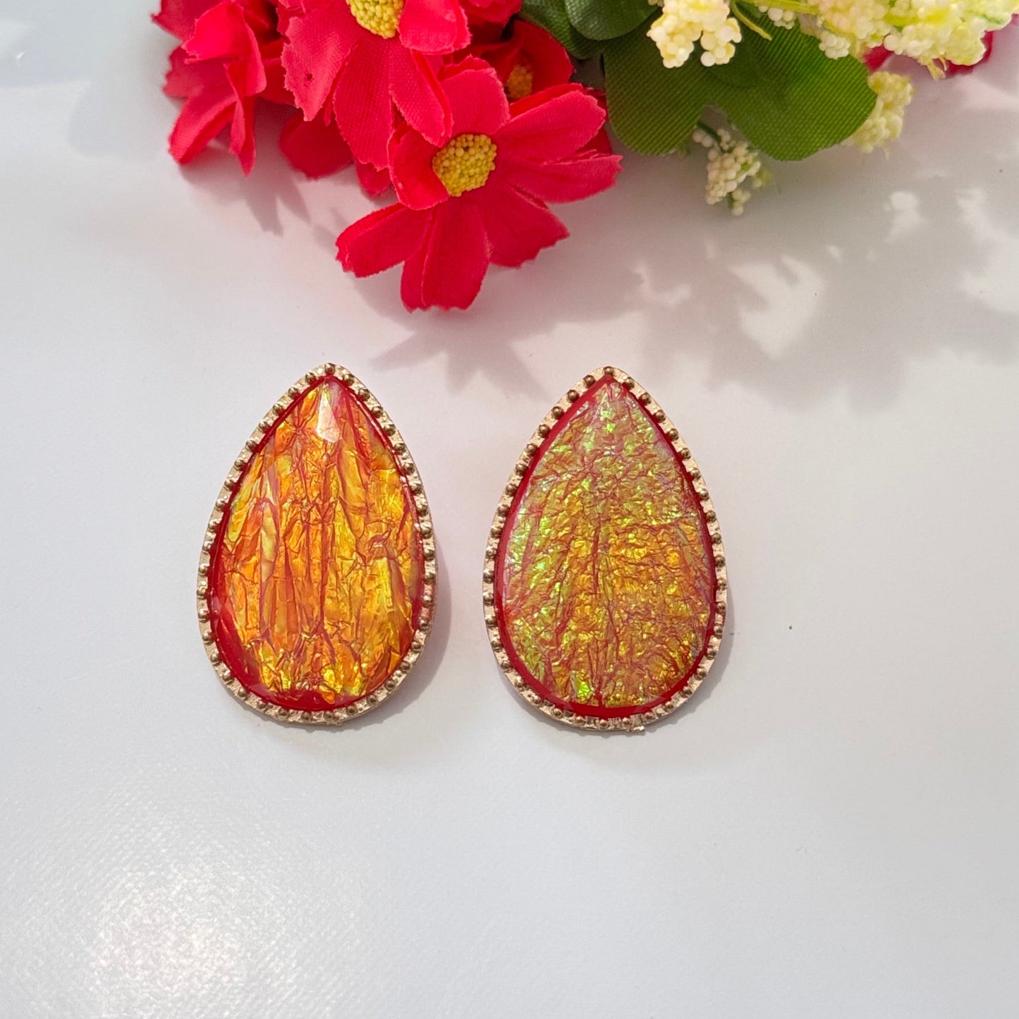 These teardrop-shaped stunners feature a mesmerizing iridescent design that shimmers with golden-orange and crimson undertones, encased in a delicate, sparkling border for a touch of elegance. The unique crinkled texture creates a captivating play of light, ensuring that these earrings stand out in any setting. 