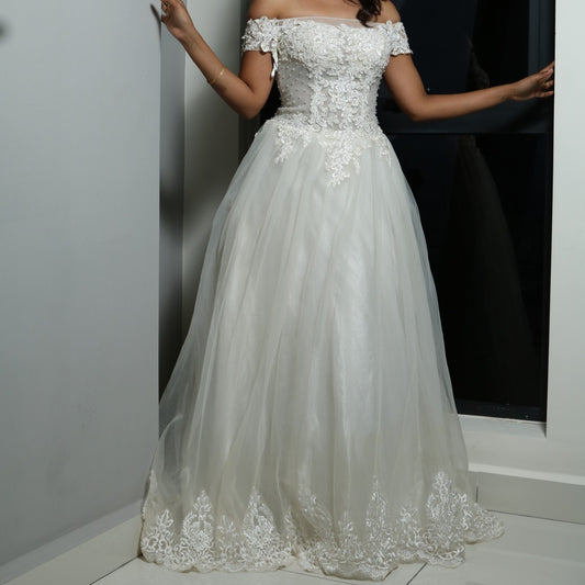 This dress is a stunning off-shoulder bridal gown with intricate lace detailing and a flowing tulle skirt. The bodice features delicate floral appliqués that add a touch of elegance, while the off-shoulder design enhances its romantic appeal. The lightweight fabric and airy structure make it an excellent choice for an outdoor or beach wedding, offering both comfort and grace. 