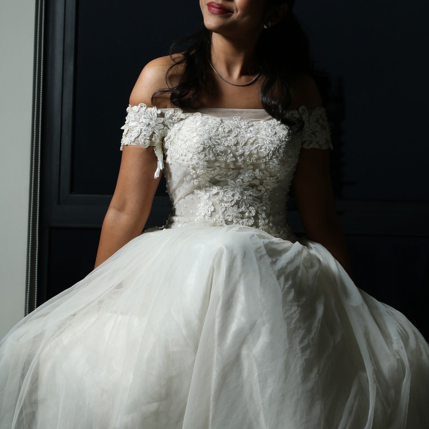 This dress is a stunning off-shoulder bridal gown with intricate lace detailing and a flowing tulle skirt. The bodice features delicate floral appliqués that add a touch of elegance, while the off-shoulder design enhances its romantic appeal. The lightweight fabric and airy structure make it an excellent choice for an outdoor or beach wedding, offering both comfort and grace. 