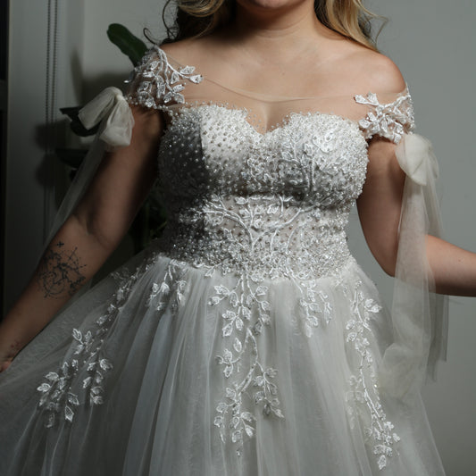 This is a light, ethereal wedding dress featuring delicate floral embroidery and beadwork. The gown has an off-the-shoulder design with sheer tulle straps tied into bows, adding a romantic touch. The bodice is adorned with intricate pearl and lace detailing, while the flowing tulle skirt is embellished with embroidered leaves, creating an elegant and whimsical look.