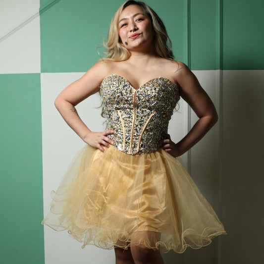 This is a glamorous party dress featuring a strapless sweetheart neckline and a fitted, heavily embellished bodice adorned with sparkling details. The structured corset-style bodice accentuates the waist, while the voluminous gold tulle skirt adds a playful and elegant touch. Perfect for special occasions, this dress combines sophistication with a fun and festive vibe.
