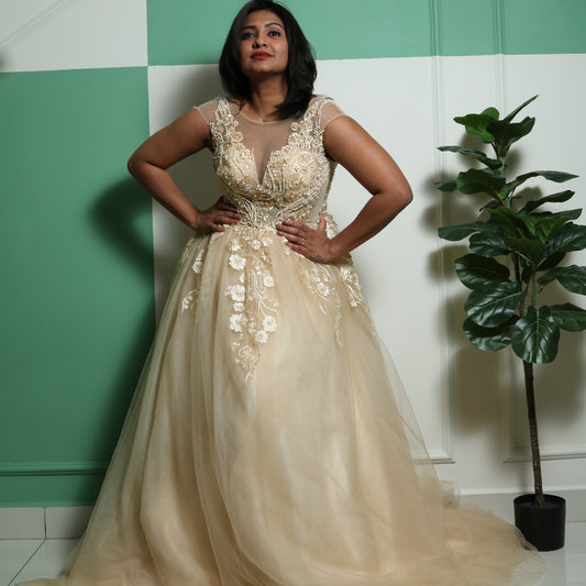 The dress in the image is a floor-length gown in a soft champagne or gold color. It features a fitted bodice with intricate floral embroidery and beadwork, extending into delicate cap sleeves. The gown has a sheer illusion neckline, adding elegance and sophistication. The voluminous tulle skirt flows gracefully, creating a princess-like silhouette. This dress is perfect for formal events, weddings, or special occasions.