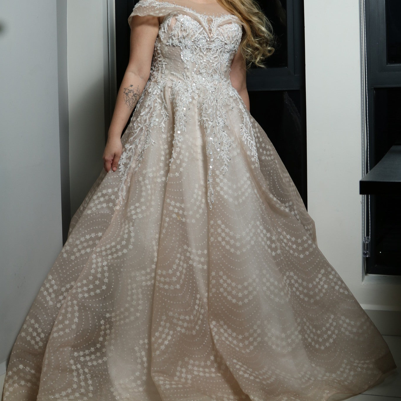 The dress in the image is an elegant, off-the-shoulder ball gown with intricate lace and bead detailing. The bodice features delicate embroidery that extends into the voluminous skirt, which is adorned with a subtle pattern of sparkling embellishments. The gown has a structured, layered design, making it visually striking but also quite heavy. The soft champagne or blush tone adds a romantic and timeless touch to the overall look.