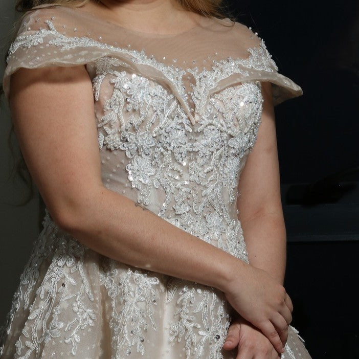 The dress in the image is an elegant, off-the-shoulder ball gown with intricate lace and bead detailing. The bodice features delicate embroidery that extends into the voluminous skirt, which is adorned with a subtle pattern of sparkling embellishments. The gown has a structured, layered design, making it visually striking but also quite heavy. The soft champagne or blush tone adds a romantic and timeless touch to the overall look.