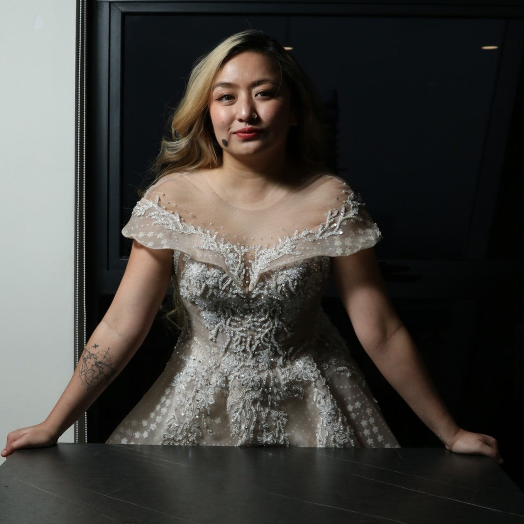 The dress in the image is an elegant, off-the-shoulder ball gown with intricate lace and bead detailing. The bodice features delicate embroidery that extends into the voluminous skirt, which is adorned with a subtle pattern of sparkling embellishments. The gown has a structured, layered design, making it visually striking but also quite heavy. The soft champagne or blush tone adds a romantic and timeless touch to the overall look.