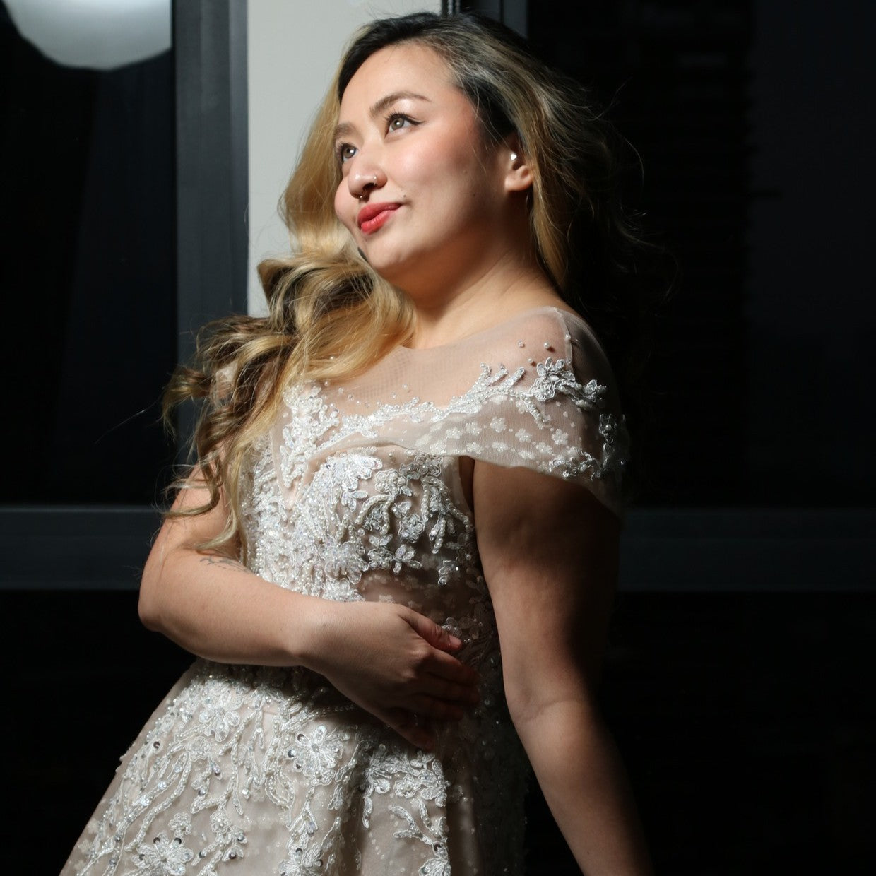 The dress in the image is an elegant, off-the-shoulder ball gown with intricate lace and bead detailing. The bodice features delicate embroidery that extends into the voluminous skirt, which is adorned with a subtle pattern of sparkling embellishments. The gown has a structured, layered design, making it visually striking but also quite heavy. The soft champagne or blush tone adds a romantic and timeless touch to the overall look.
