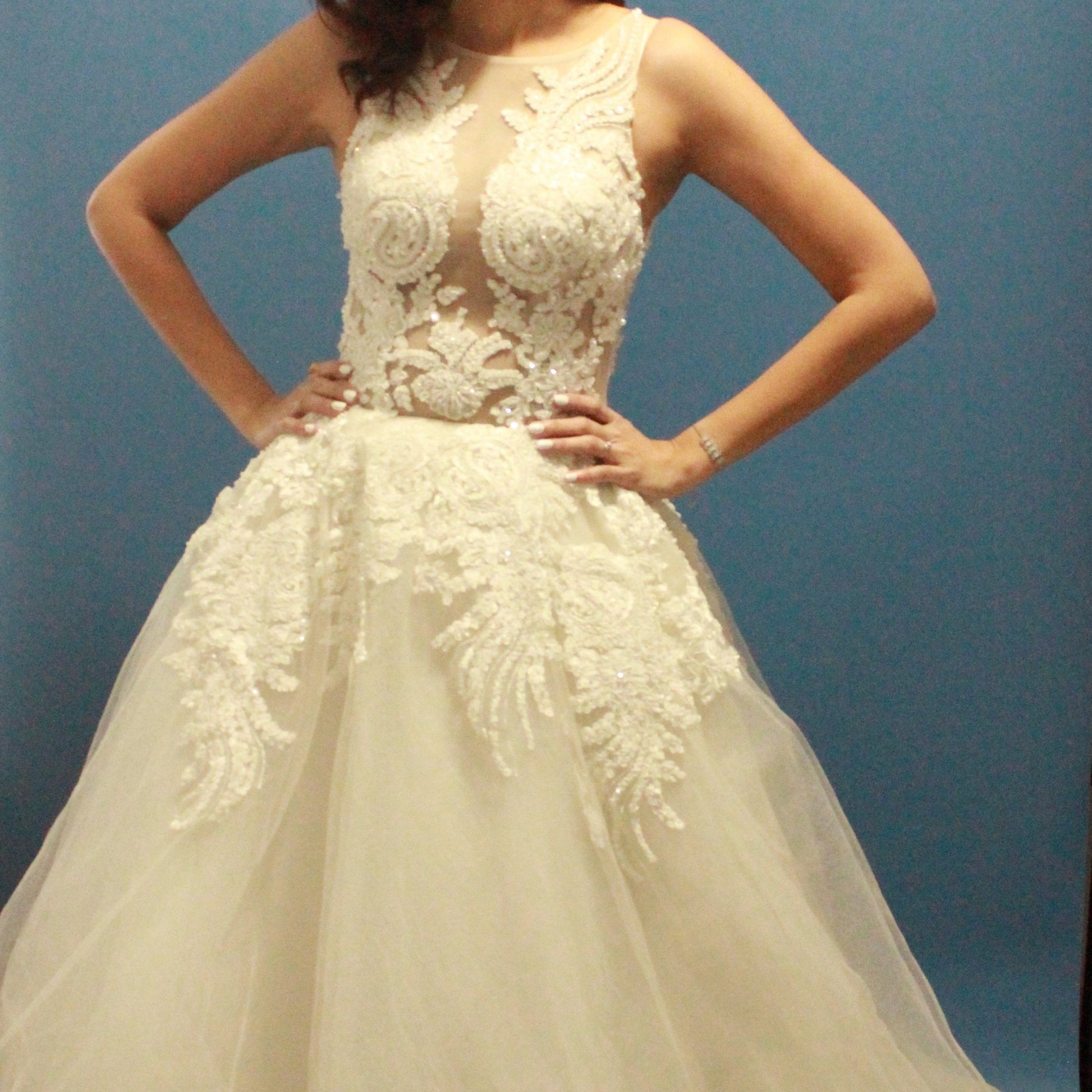 This wedding gown is a breathtaking blend of elegance and drama. The bodice features intricate lace appliqué and shimmering beadwork on a sheer illusion panel that tastefully reveals the décolletage, creating a striking yet sophisticated look. The voluminous skirt, adorned with cascading lace details and layers of soft tulle, adds to the gown’s regal presence. 