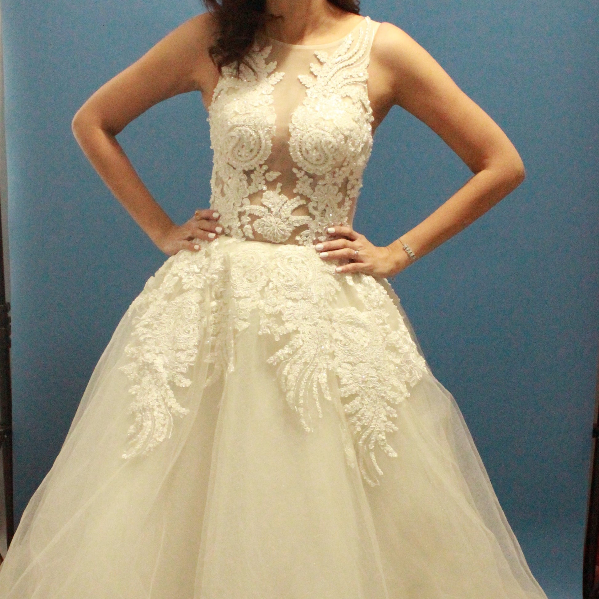 This wedding gown is a breathtaking blend of elegance and drama. The bodice features intricate lace appliqué and shimmering beadwork on a sheer illusion panel that tastefully reveals the décolletage, creating a striking yet sophisticated look. The voluminous skirt, adorned with cascading lace details and layers of soft tulle, adds to the gown’s regal presence. 