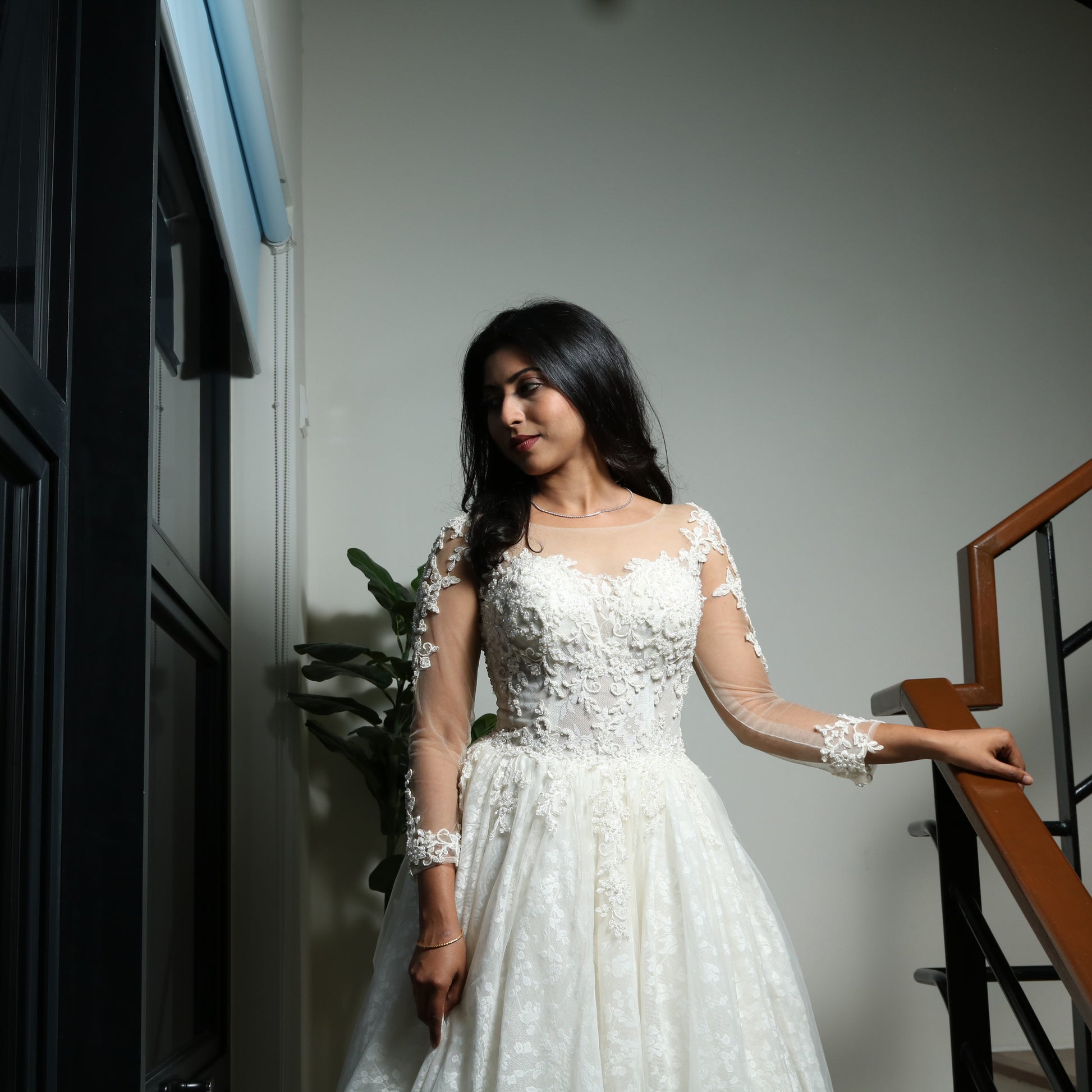This wedding dress is an elegant A-line gown featuring a sheer illusion neckline, long lace-embellished sleeves, and a fitted bodice adorned with delicate floral lace appliqués. The voluminous skirt, crafted from soft tulle with lace detailing, adds a romantic and timeless touch, making it a perfect choice for a classic bridal look.