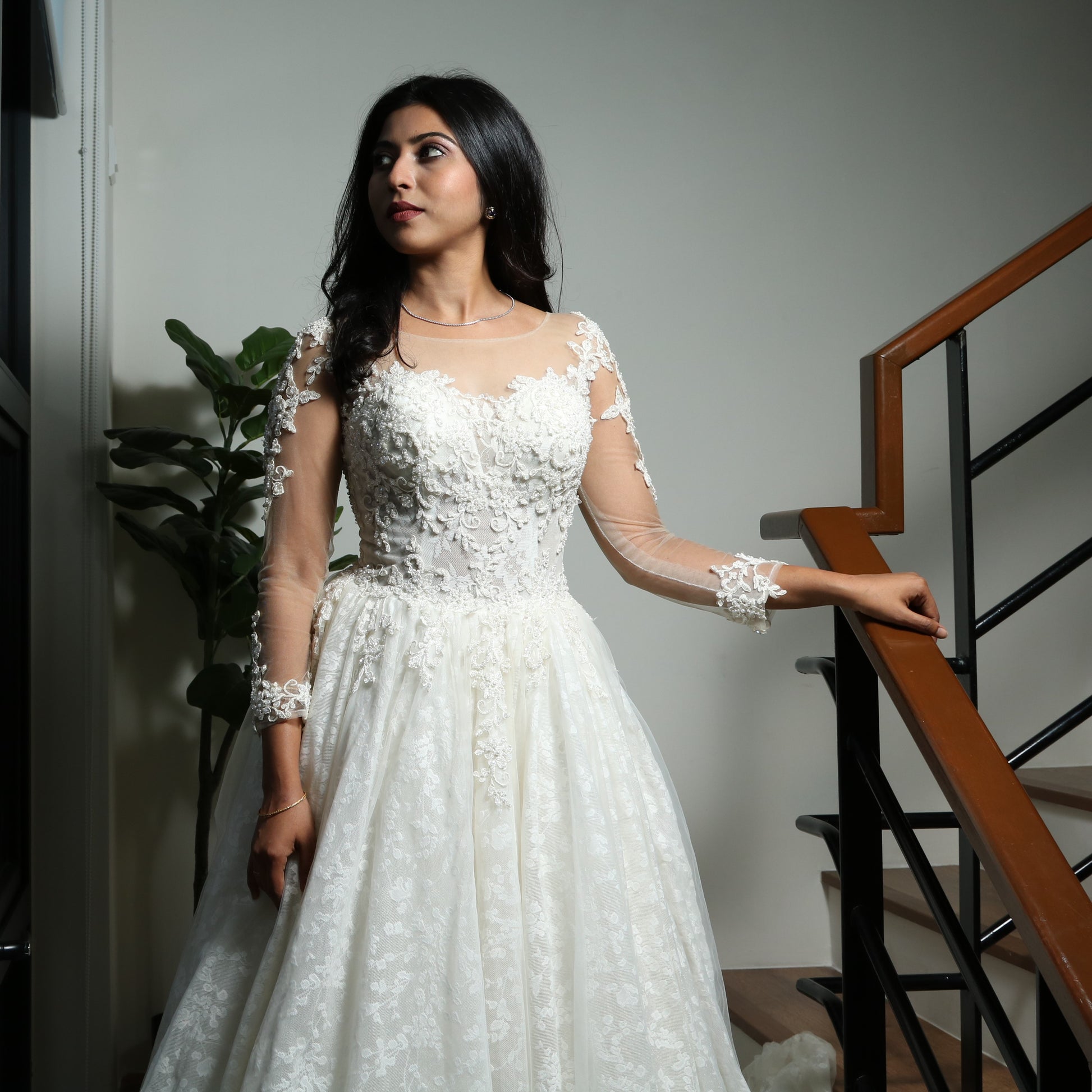 This wedding dress is an elegant A-line gown featuring a sheer illusion neckline, long lace-embellished sleeves, and a fitted bodice adorned with delicate floral lace appliqués. The voluminous skirt, crafted from soft tulle with lace detailing, adds a romantic and timeless touch, making it a perfect choice for a classic bridal look.