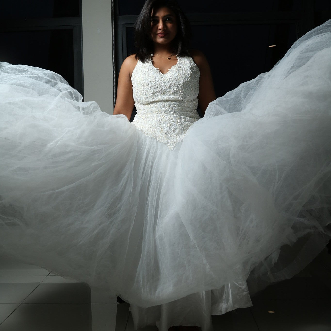 This dress is a stunning white gown with a voluminous tulle skirt, creating an ethereal and lightweight appearance. The bodice features intricate lace or beaded detailing with a flattering V-neckline and sleeveless design. The skirt is made of multiple layers of airy tulle, giving it a dreamy, floating effect. The overall look is elegant, romantic, and light, making it a perfect choice for a formal event such as a wedding or a gala.