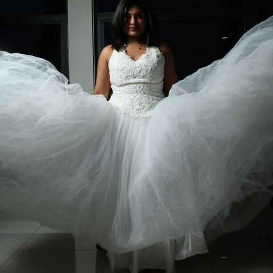 This dress is a stunning white gown with a voluminous tulle skirt, creating an ethereal and lightweight appearance. The bodice features intricate lace or beaded detailing with a flattering V-neckline and sleeveless design. The skirt is made of multiple layers of airy tulle, giving it a dreamy, floating effect. The overall look is elegant, romantic, and light, making it a perfect choice for a formal event such as a wedding or a gala.