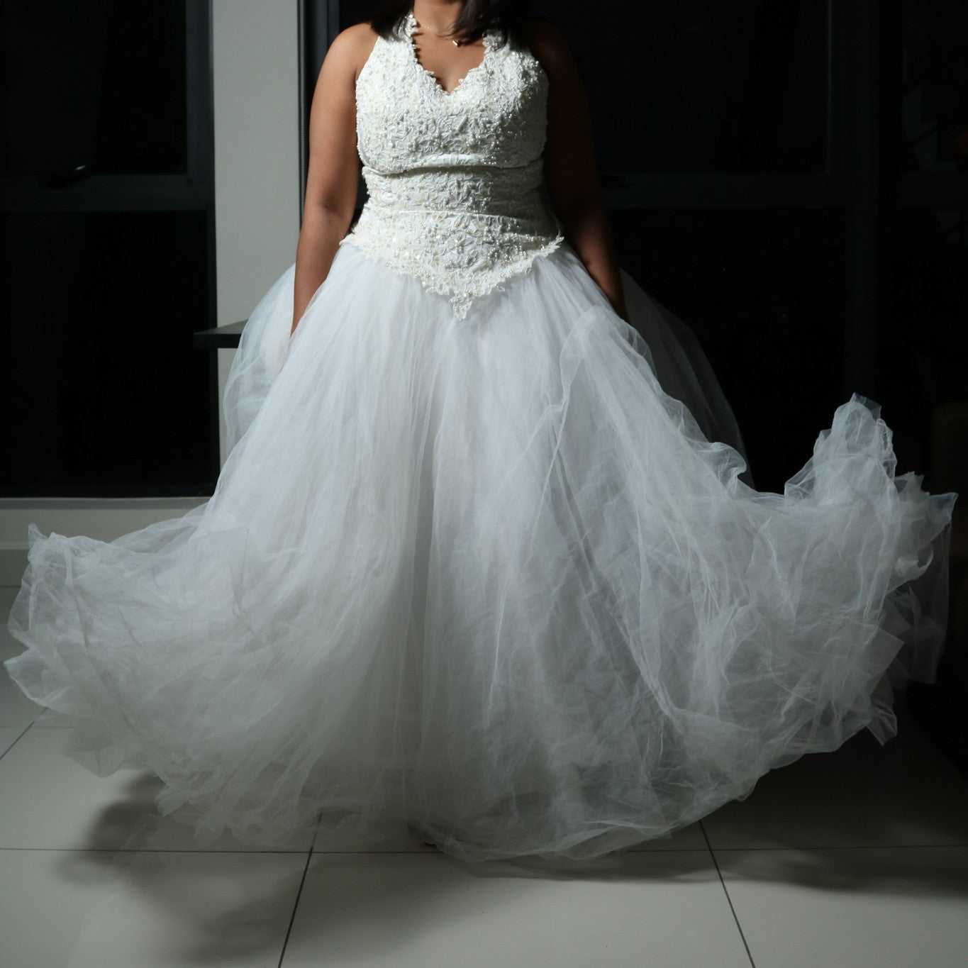 This dress is a stunning white gown with a voluminous tulle skirt, creating an ethereal and lightweight appearance. The bodice features intricate lace or beaded detailing with a flattering V-neckline and sleeveless design. The skirt is made of multiple layers of airy tulle, giving it a dreamy, floating effect. The overall look is elegant, romantic, and light, making it a perfect choice for a formal event such as a wedding or a gala.