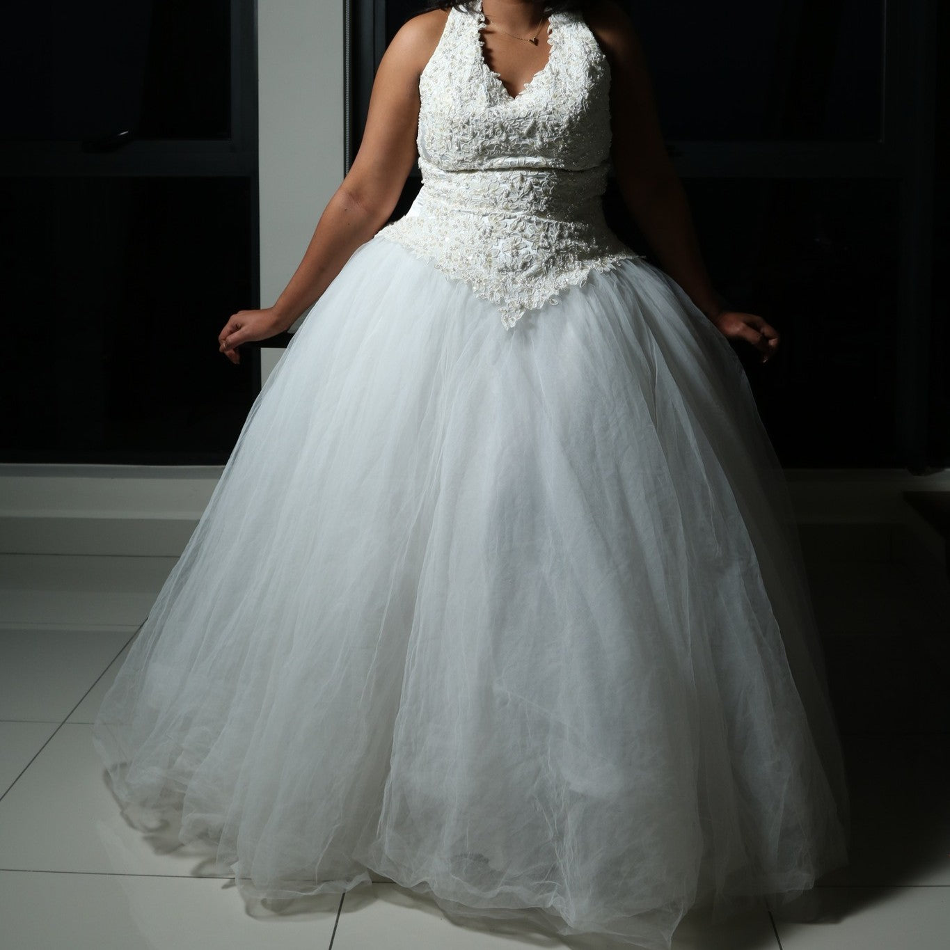 This dress is a stunning white gown with a voluminous tulle skirt, creating an ethereal and lightweight appearance. The bodice features intricate lace or beaded detailing with a flattering V-neckline and sleeveless design. The skirt is made of multiple layers of airy tulle, giving it a dreamy, floating effect. The overall look is elegant, romantic, and light, making it a perfect choice for a formal event such as a wedding or a gala.
