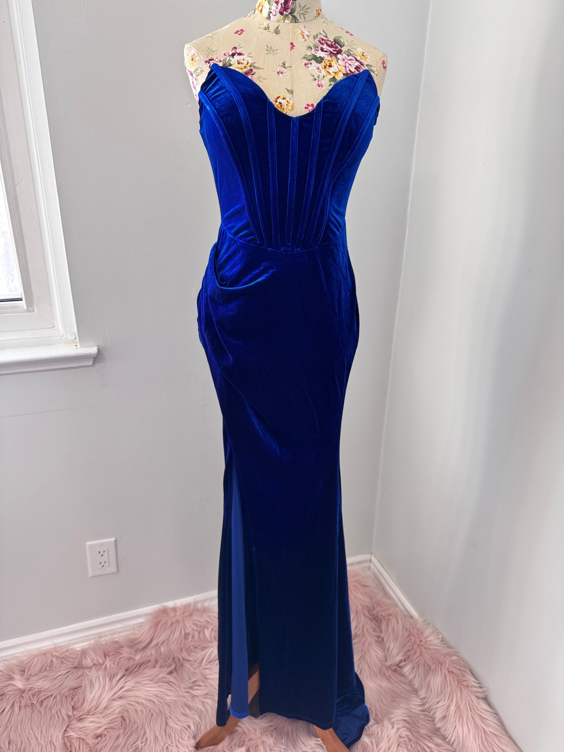 Step into timeless elegance with this breathtaking previously cherished royal blue velvet evening gown, designed to turn heads at any event. Crafted from rich, ultra-soft velvet, this figure-hugging masterpiece features a structured corset bodice with delicate pleated detailing that accentuates your silhouette. The sweetheart neckline adds a touch of romance, while the flowing skirt with a sultry thigh-high slit creates the perfect balance of sophistication and allure. In perfect condition.